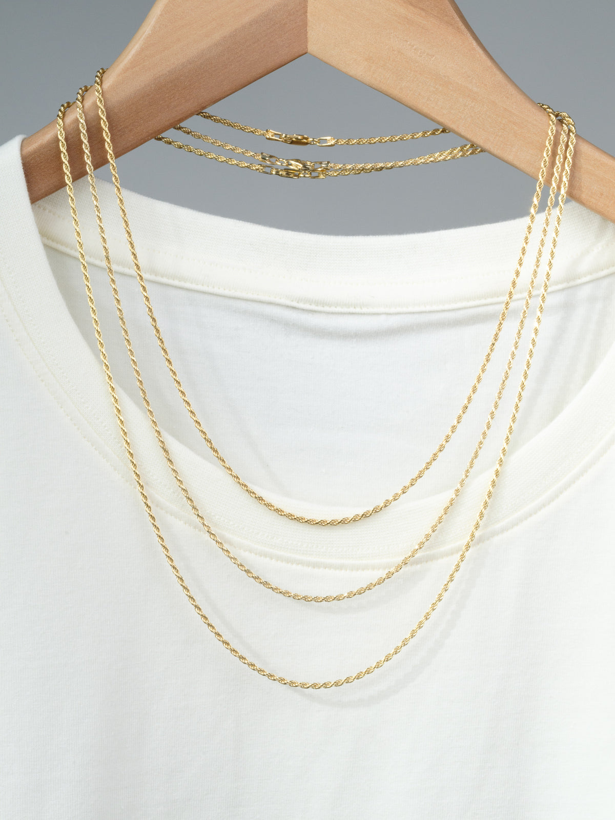 The Orion 40 Rope Chain Necklace by ID Menswear is draped over a white t-shirt on a wooden hanger, featuring two thin chains, all displayed against a plain gray background.