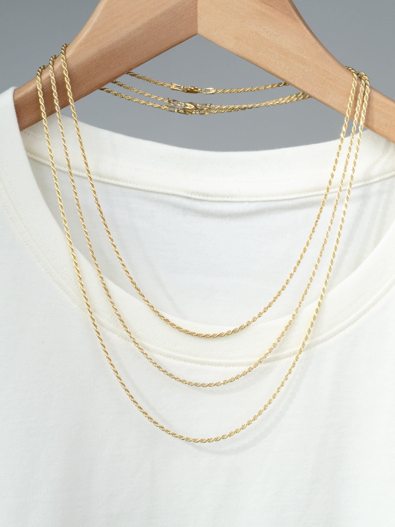 The Orion 40 Rope Chain Necklace by ID Menswear is draped over a white t-shirt on a wooden hanger, featuring two thin chains, all displayed against a plain gray background.