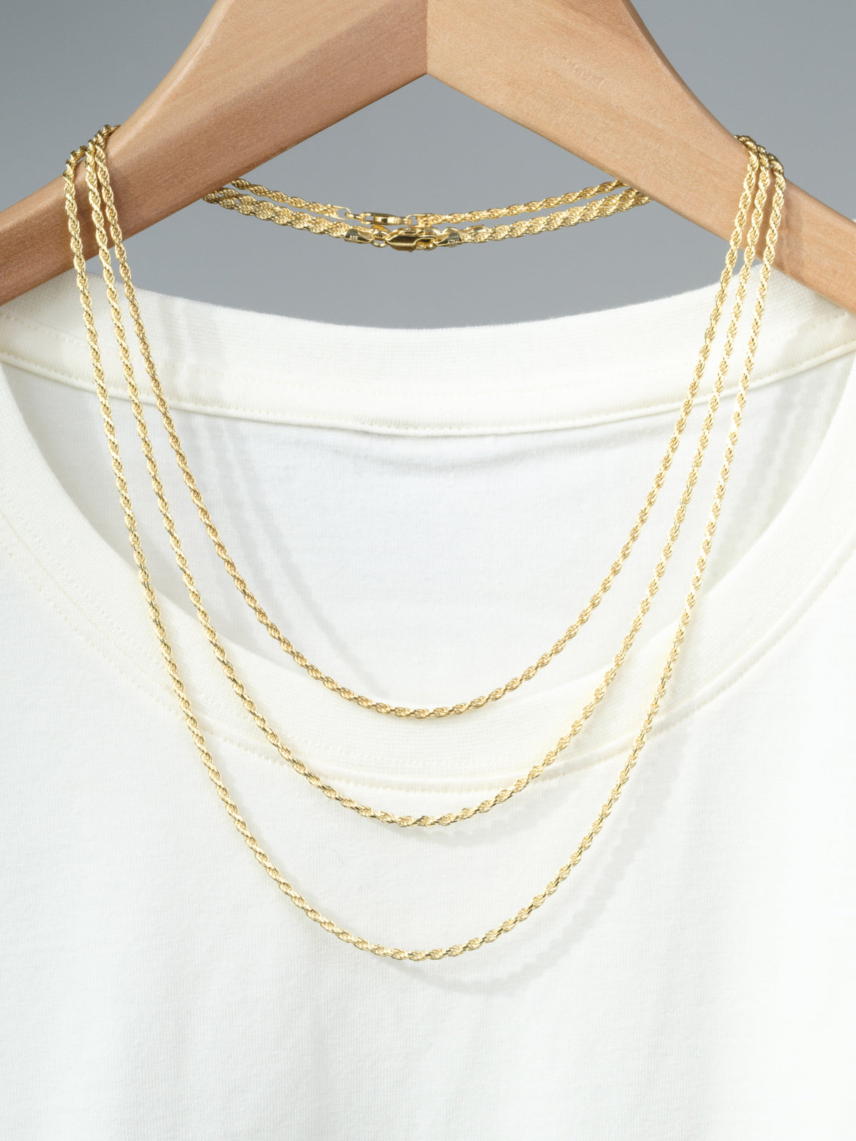 A white T-shirt on a wooden hanger displays the ID Menswear Orion 50 Rope Chain Necklace, draped elegantly over the shirt’s neckline against a plain gray background.