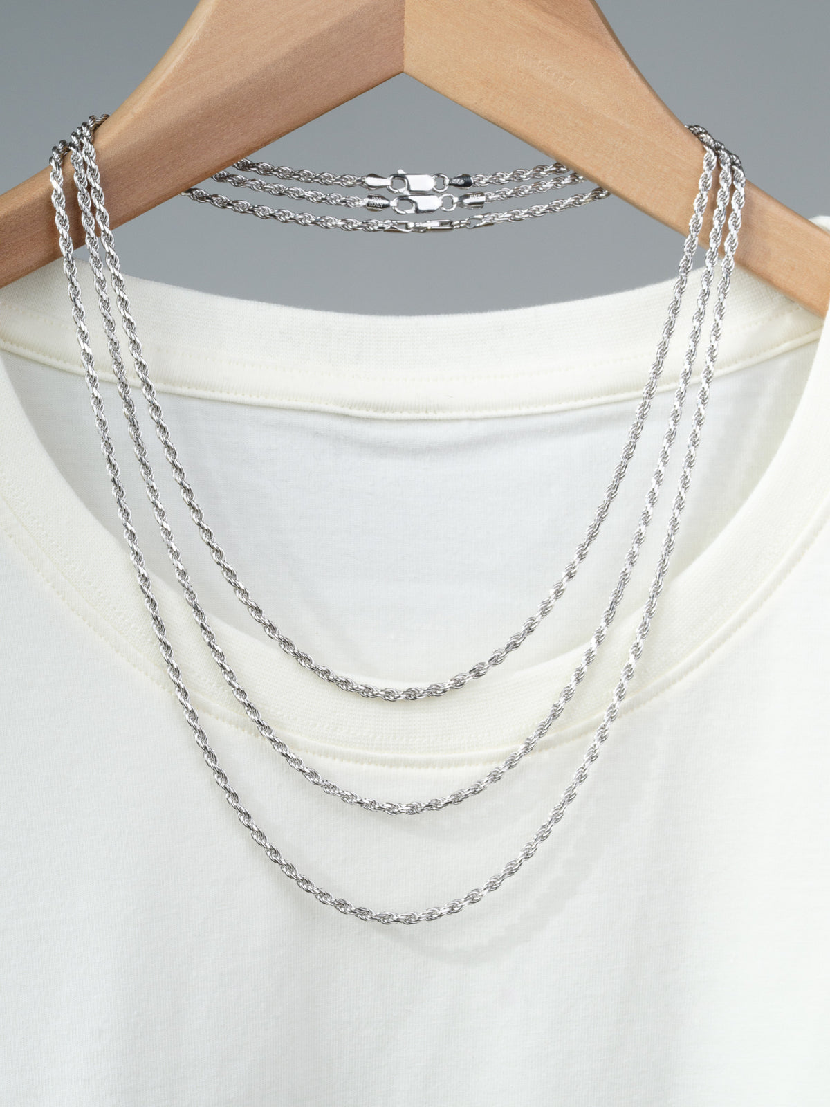 A white T-shirt on a wooden hanger displays three layered Orion 60 Rope Chain Necklaces by ID Menswear, including a 5mm Sterling Silver option. Each piece’s Italian craftsmanship stands out against the plain gray background.