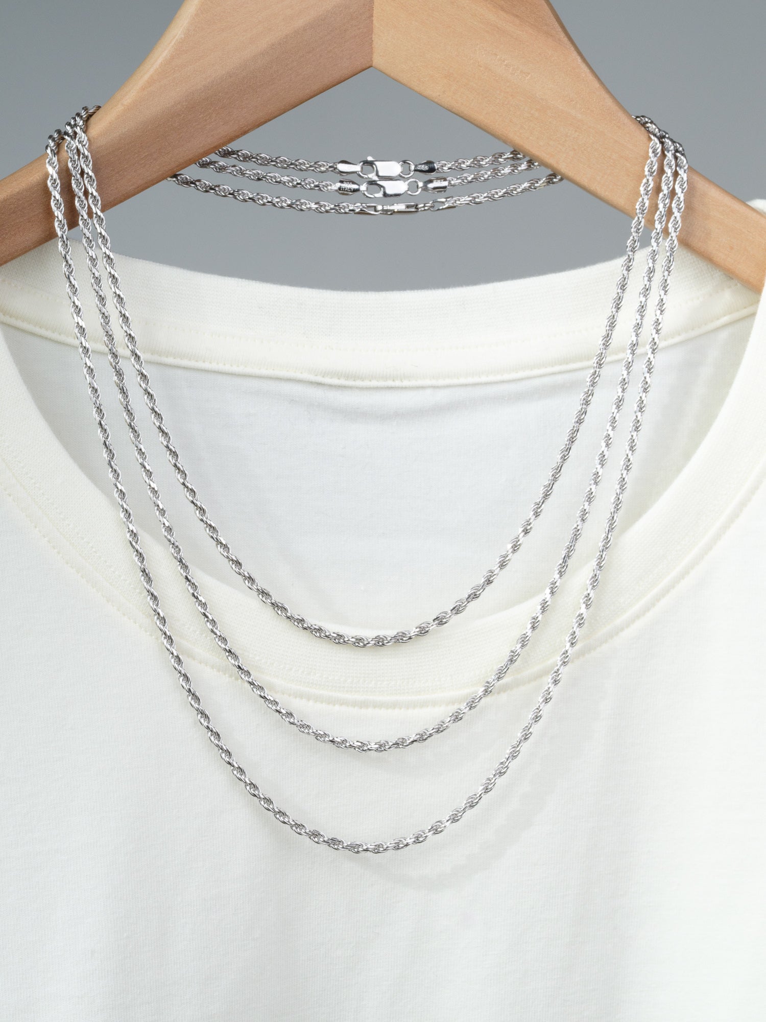 A white T-shirt on a wooden hanger displays three layered Orion 60 Rope Chain Necklaces by ID Menswear, including a 5mm Sterling Silver option. Each piece’s Italian craftsmanship stands out against the plain gray background.