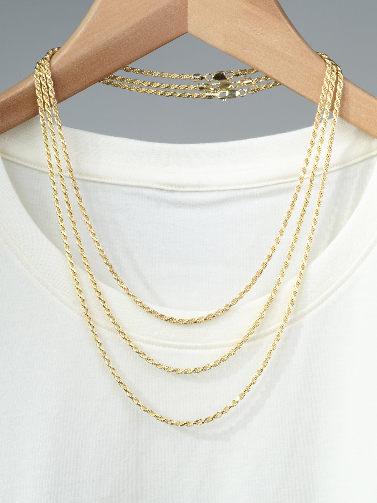 An Orion 60 Rope Chain Necklace by ID Menswear drapes over a white T-shirt on a wooden hanger, alongside two gold rope chains of varying lengths, all set against a neutral gray background.