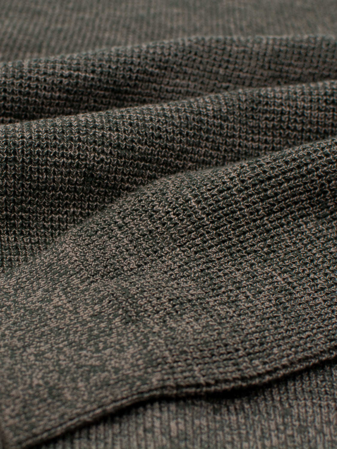 Close-up of the Oslo Waffle Knit Sweater by ID Menswear, showcasing textured gray knit fabric with a subtle checkered pattern. Reminiscent of cozy cotton sweaters, the soft material is slightly folded to highlight its woven detail, making it ideal for stylish yet comfortable casual wear.