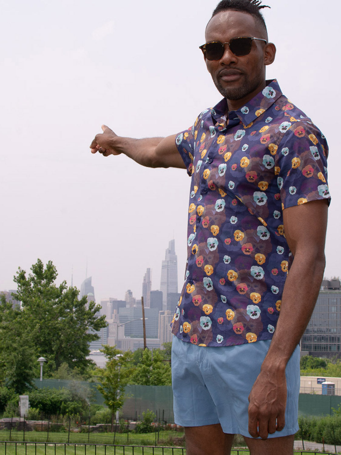 A man in ID Menswear’s Oyogu Quick Dry 4 Way Stretch Swim Trunk, sunglasses, and a colorful shirt stands outdoors pointing at a distant city skyline through haze, with trees and buildings in the foreground.