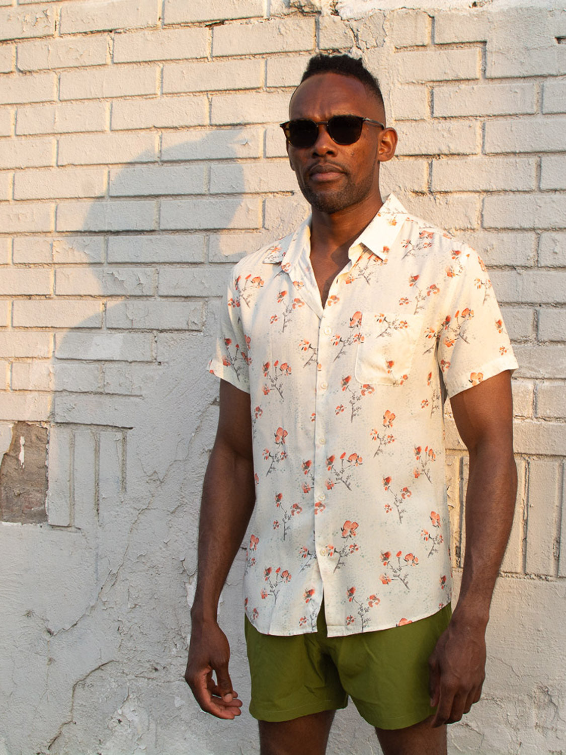 A man in sunglasses, a light floral short-sleeve shirt, and olive green ID Menswear Oyogu Quick Dry 4 Way Stretch Swim Trunk made from recycled plastic stands against a sunlit white brick wall.