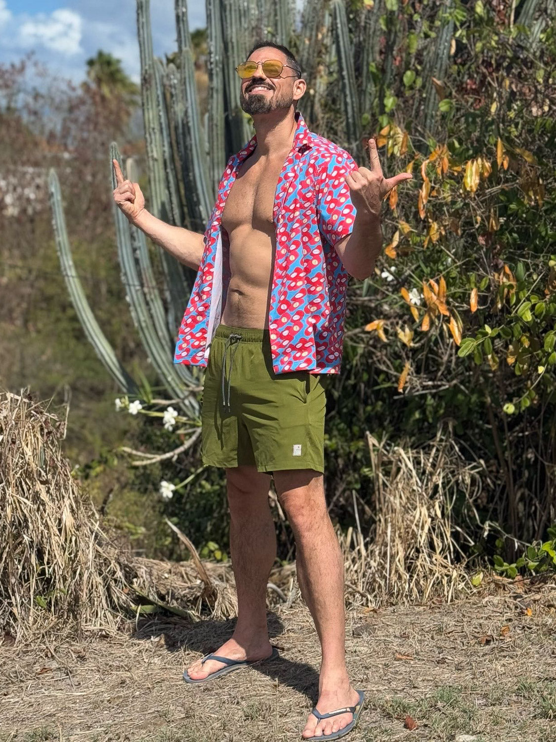 A man stands outdoors in front of cacti, smiling with arms raised. Hes wearing ID Menswears Oyogu Quick Dry 4 Way Stretch Swim Trunk—olive green shorts made from recycled plastic bottles—and sunglasses under bright sunlight.