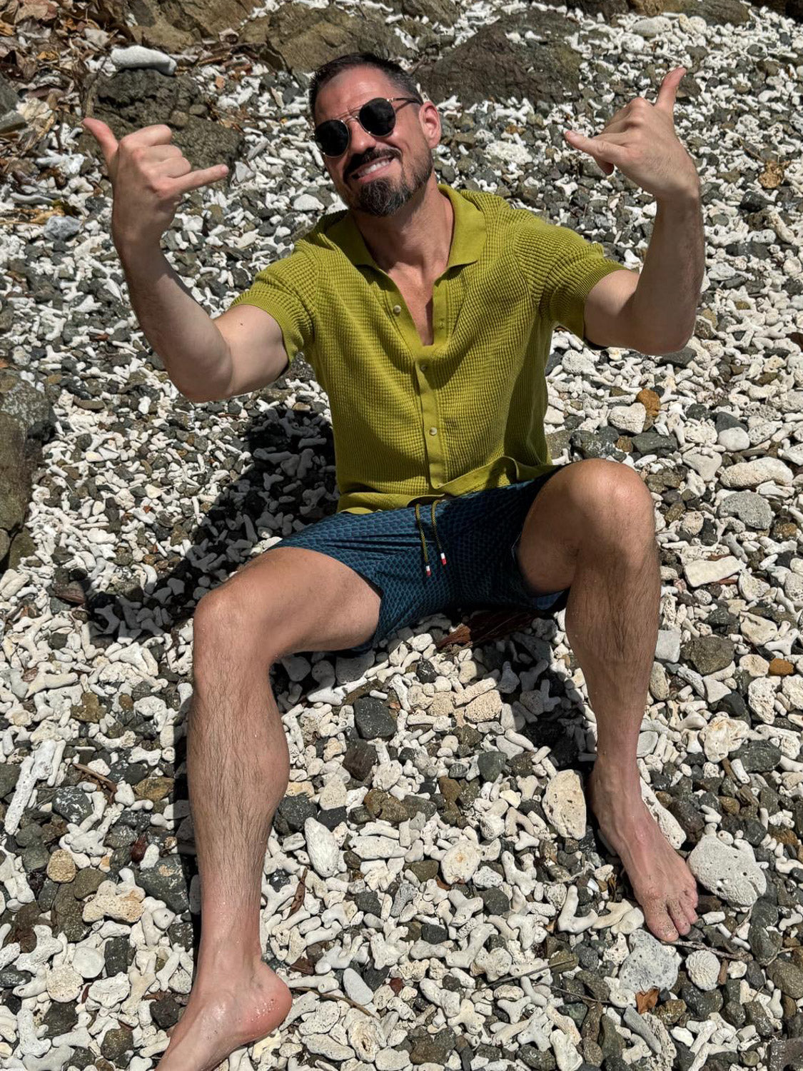 A man in ID Menswears Uroko Fast Dry 4-Way Stretch Swim Trunk sits barefoot on a rocky, sunlit beach, wearing sunglasses and a green shirt, smiling and making a shaka hand gesture with both hands.