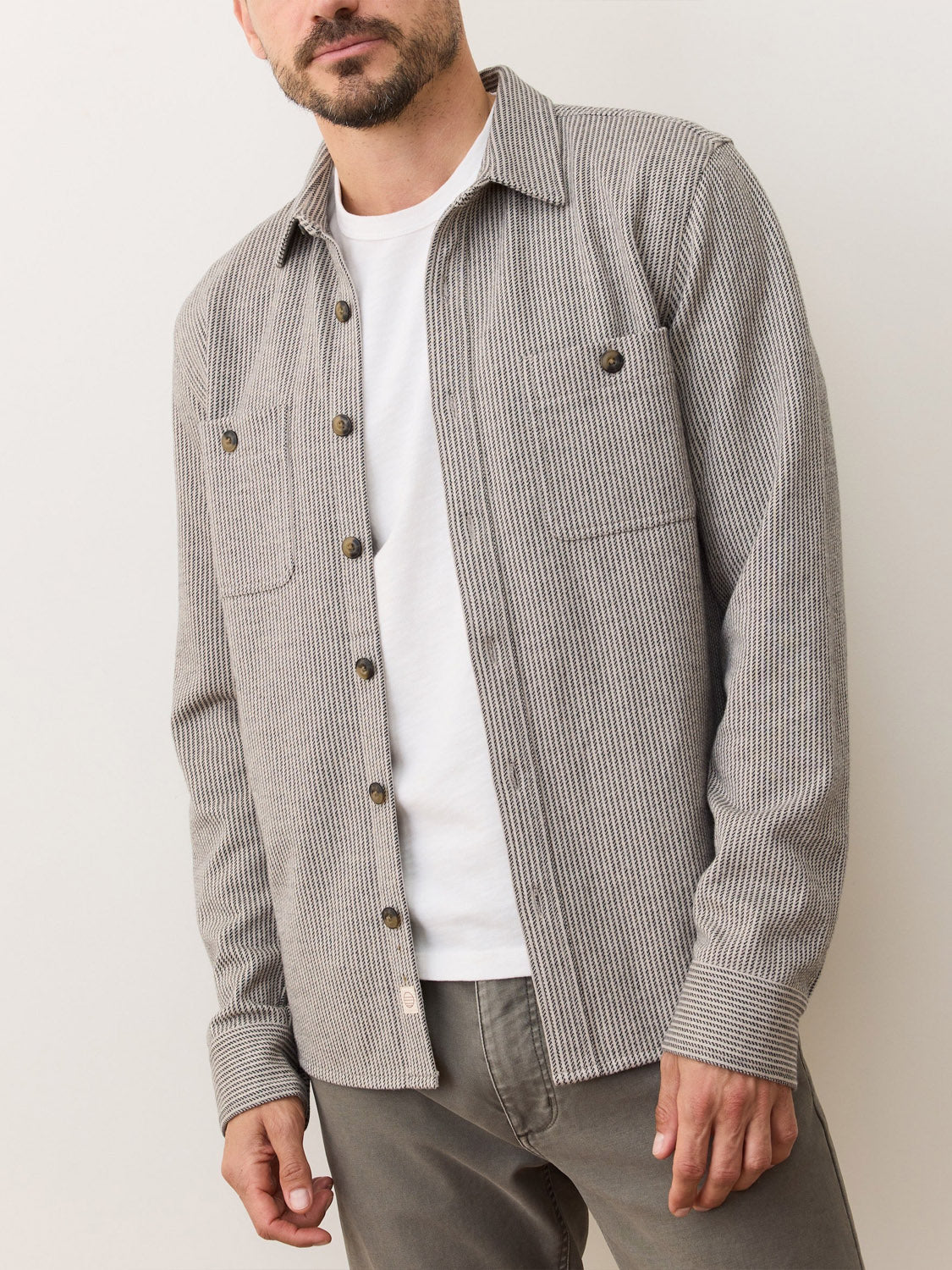 A man wearing a Marine Layer Pacific Stretch Grey Twill Overshirt over a white t-shirt and grey pants stands against a plain light background.