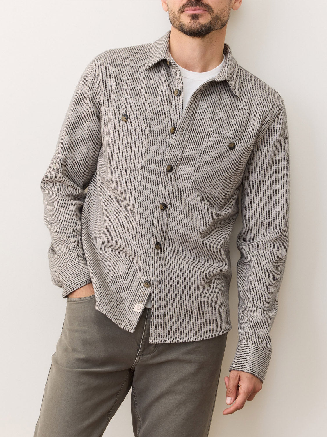 A man wearing a Marine Layer Pacific Stretch Grey Twill Overshirt over a white t-shirt and gray pants stands against a light background with one hand in his pocket.