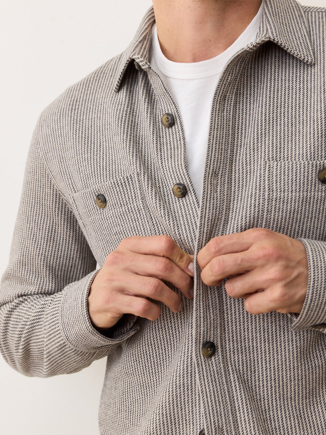 A person is buttoning a Marine Layer Pacific Stretch Grey Twill Overshirt over a white t-shirt. Only their torso and hands are visible. The grey overshirt features dark buttons and a chest pocket.