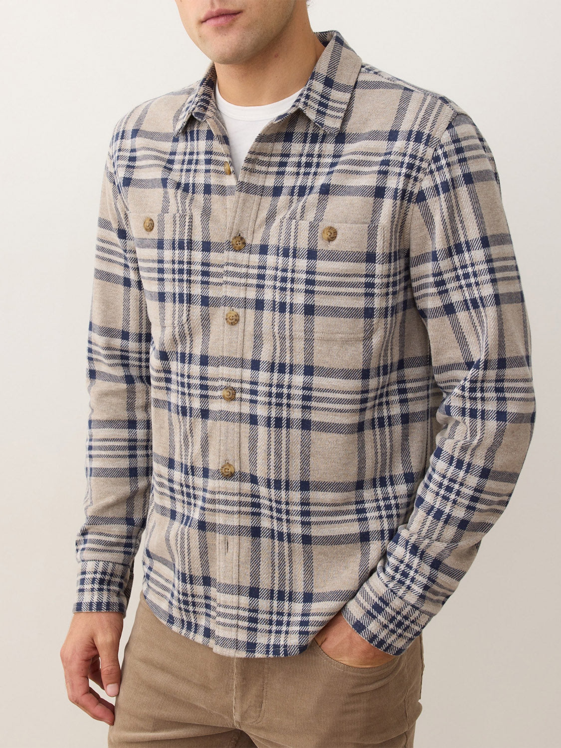 A man wearing the Marine Layer Pacific Stretch Plaid Twill Overshirt in beige and blue with front pockets, paired with light brown pants, stands against a neutral background.
