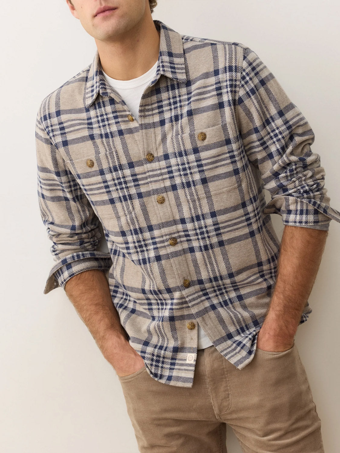 A man wears Marine Layer’s Pacific Stretch Plaid Twill Overshirt in beige and blue over a white tee with tan pants, standing hands-in-pockets against a light background.