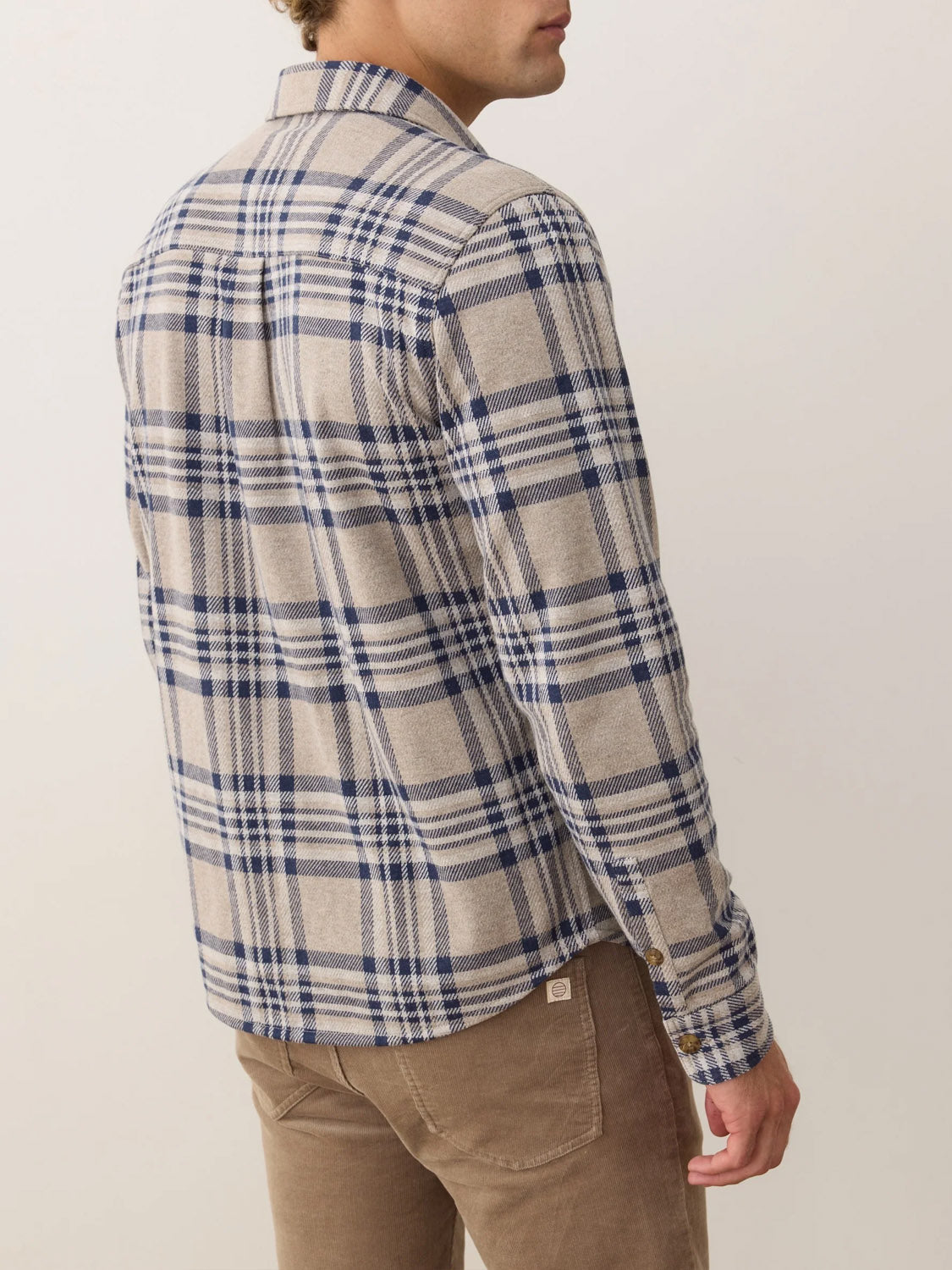 A man stands with his back turned, wearing Marine Layer’s Pacific Stretch Plaid Twill Overshirt in beige and blue plaid, paired with light brown pants against a plain, light background.