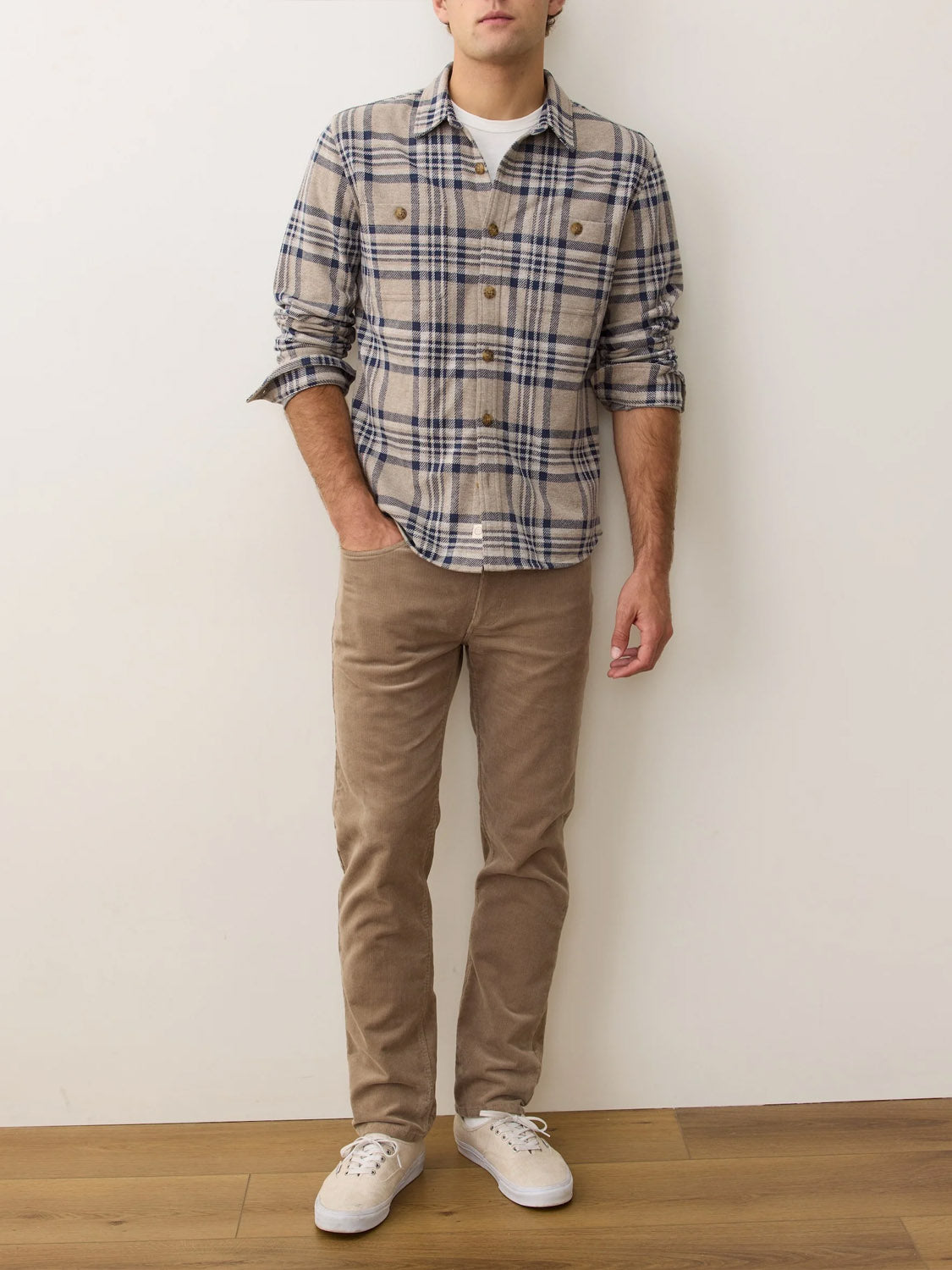 A man stands against a white wall wearing a Marine Layer Pacific Stretch Plaid Twill Overshirt in blue and beige, paired with light brown corduroy pants and white sneakers. His face is not visible.