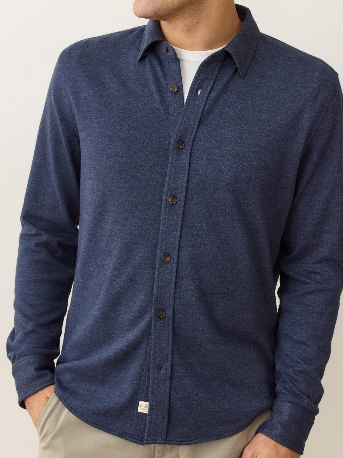 A man wears a navy Pacific Stretch Twill Shirt by Marine Layer with long sleeves and beige pants, left hand in pocket, looking slightly to the side. The soft stretch fabric enhances his comfort and style.