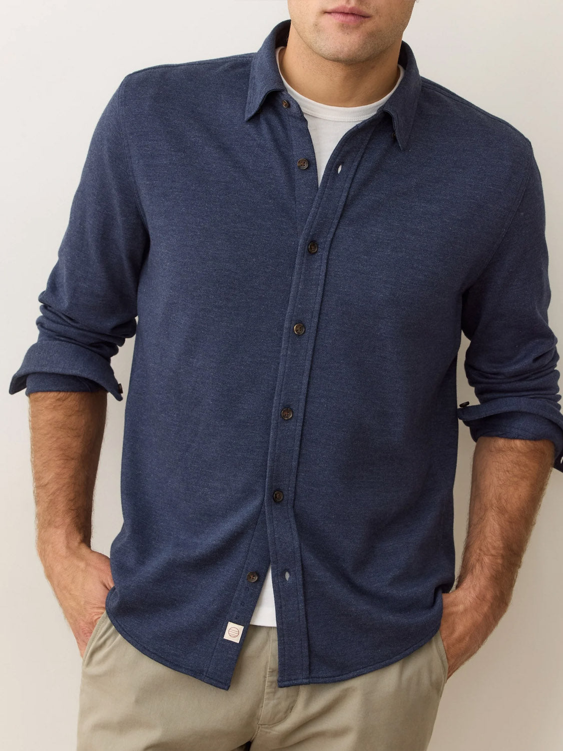A man wears a Marine Layer Pacific Stretch Twill Shirt—a dark blue men’s overshirt made from soft stretch twill—over a white tee and beige pants. With rolled-up sleeves and hands in pockets, he faces forward, his face partially out of frame.