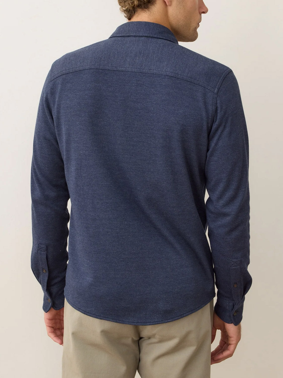 A man stands with his back to the camera against a plain, light background, wearing a long-sleeve, dark blue Marine Layer Pacific Stretch Twill Shirt and light beige pants, highlighting the comfort of its soft stretch fabric.
