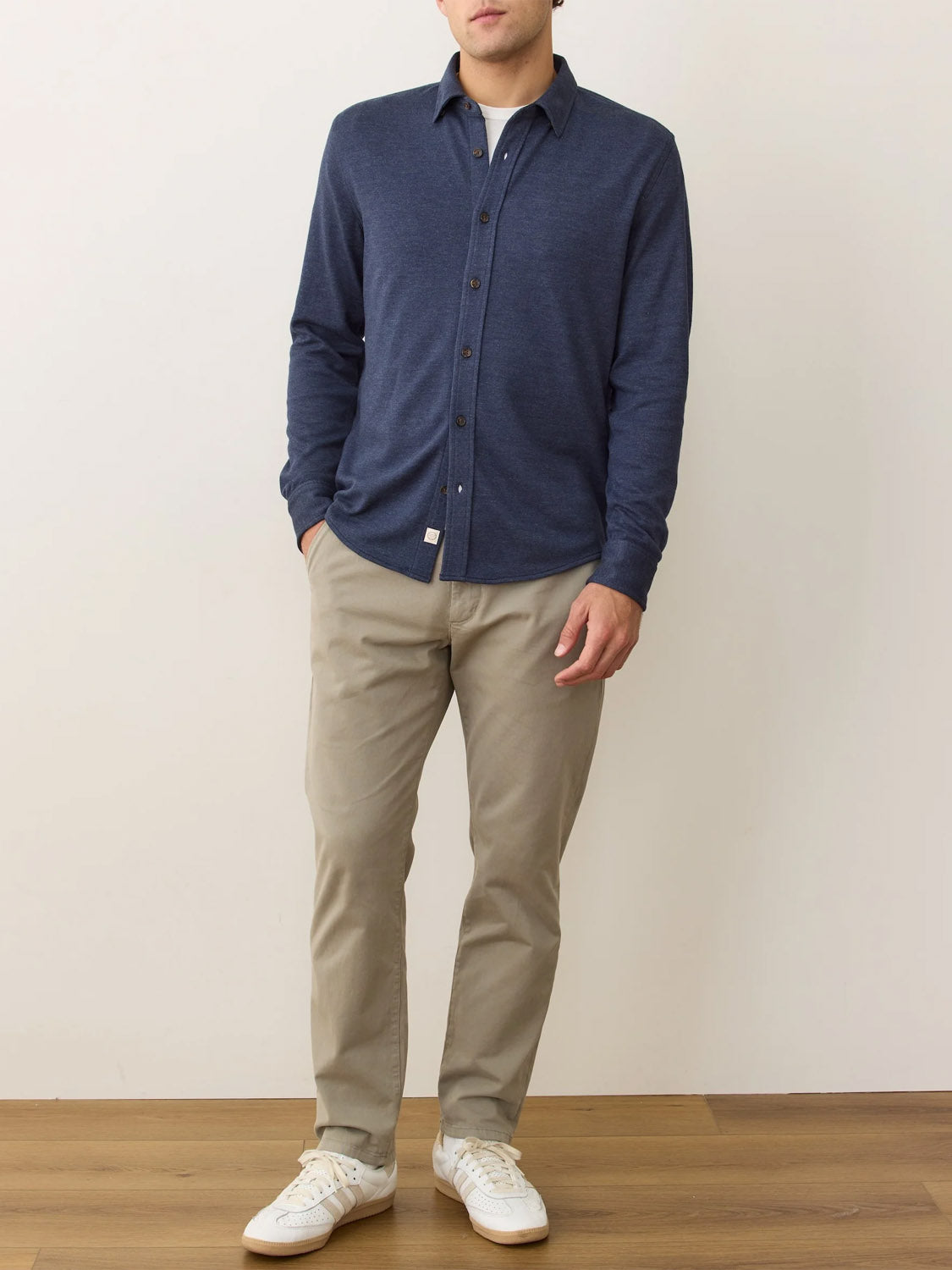 A man stands against a plain wall, wearing a Marine Layer Pacific Stretch Twill Shirt in blue, beige pants, and white sneakers. With his hands in his pockets and face partially out of frame, the shirt’s soft stretch fabric is highlighted.