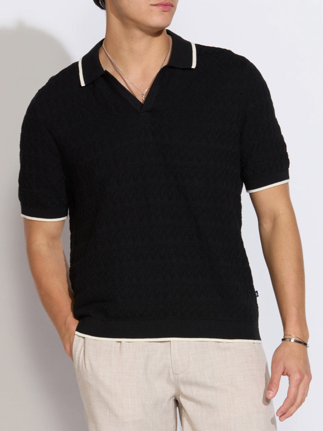 A person wearing the Civil Society Palani Johnny Collar Sweater Polo in black with white trim, made from soft cotton yarn, paired with light beige pants, poses with one hand in their pocket against a plain background.