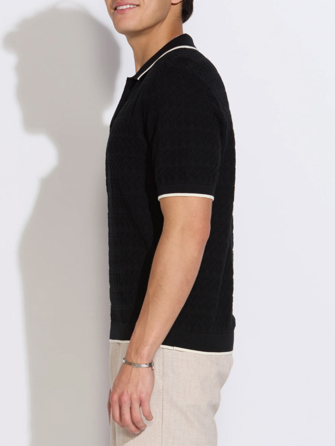 A man stands in profile against a white background, wearing the Palani Johnny Collar Sweater Polo by Civil Society in black cotton yarn with white trim and light beige pants. His face is partially out of frame, and he also wears a silver bracelet.