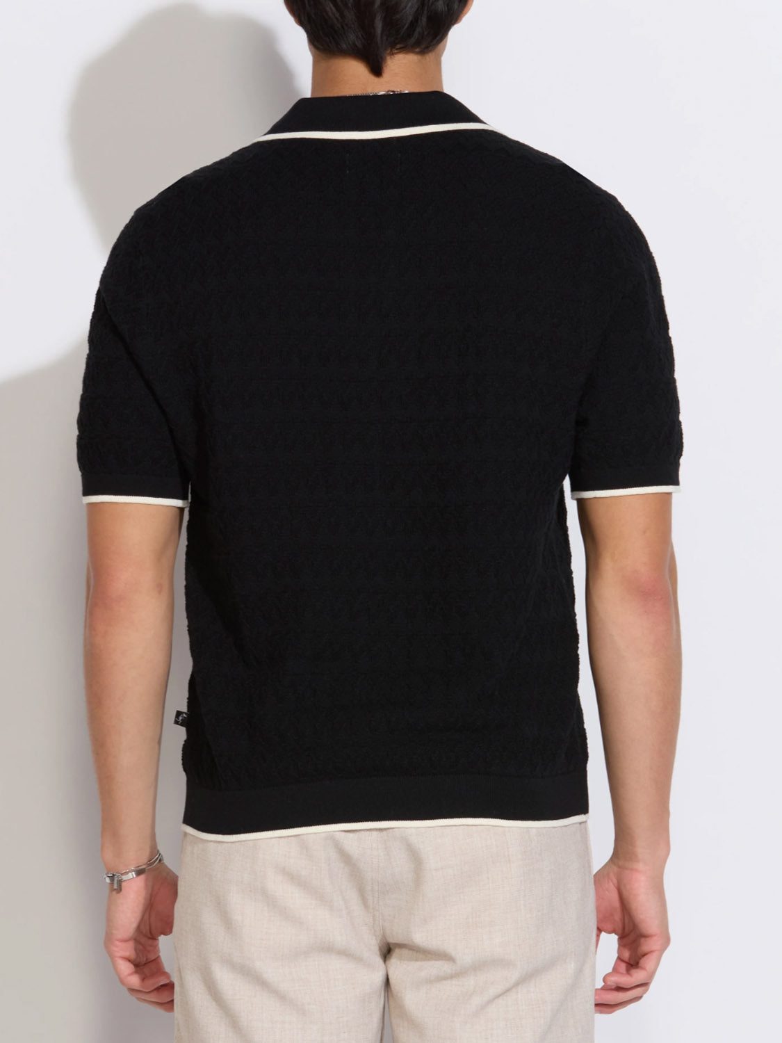 A person is shown from the back wearing the Civil Society Palani Johnny Collar Sweater Polo in black with white trim and light beige pants, against a plain light-colored background.
