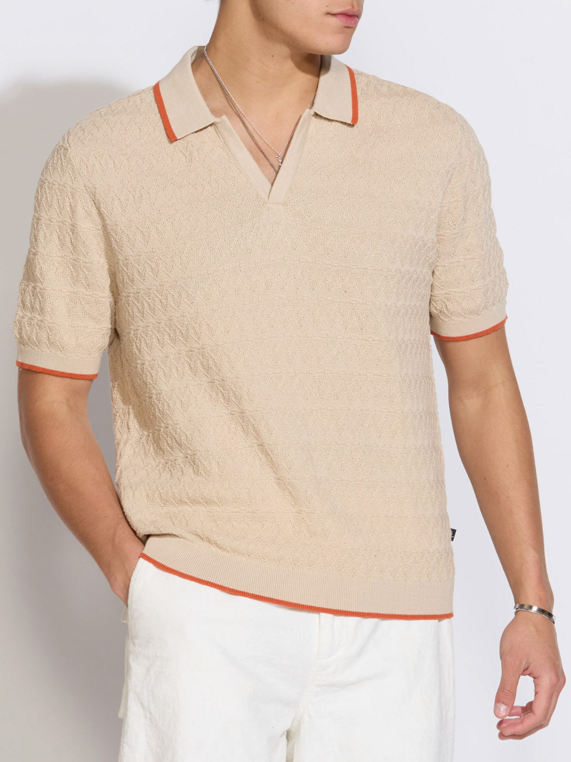 A person stands against a plain light background wearing the Civil Society Palani Johnny Collar Sweater Polo in beige with orange trim, styled in a relaxed fit, paired with white pants and a silver necklace.