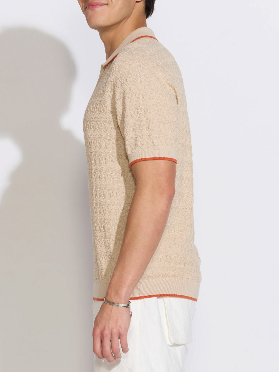 A person stands in profile against a white background, wearing the Civil Society Palani Johnny Collar Sweater Polo in light beige with orange trim and white pants. They have short hair and wear a silver bracelet on their wrist.