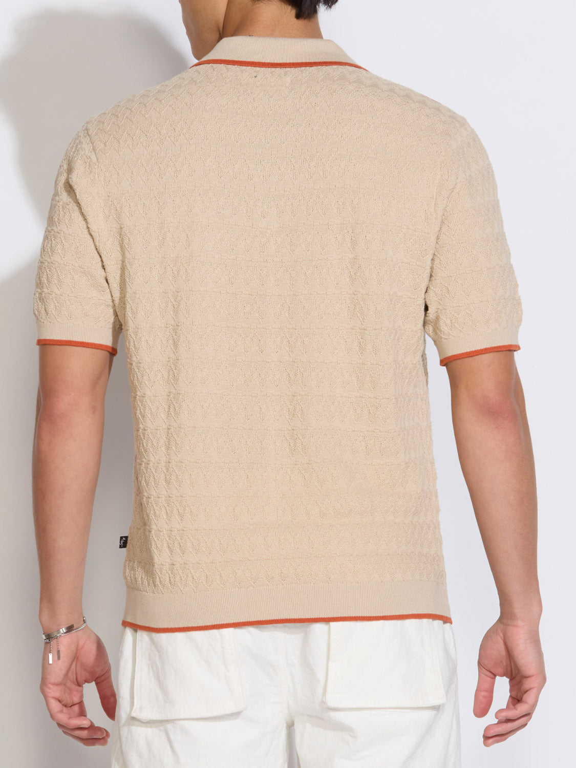 Someone is seen from behind wearing Civil Society’s Palani Johnny Collar Sweater Polo in beige with orange trim on the collar and sleeves, styled with white shorts. The polo features a relaxed fit and short sleeves.