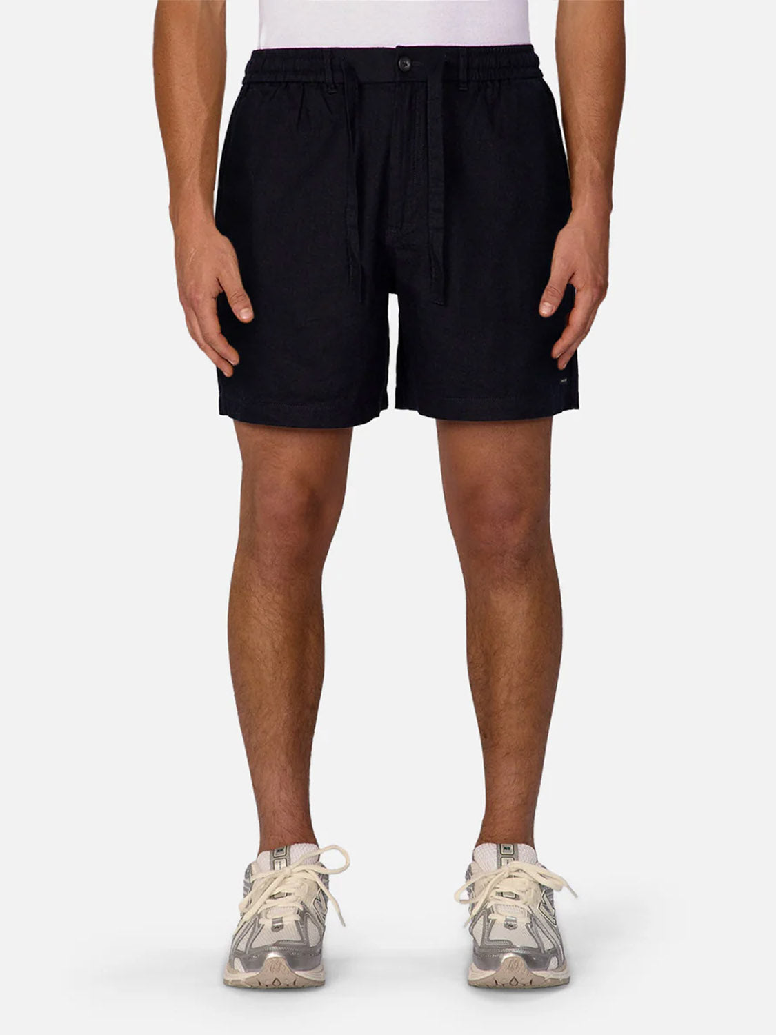 A person stands facing forward wearing Industrie Palma Linen Short in black with a drawstring and light gray patterned sneakers. Their hands rest at their sides against a plain, light-colored background.