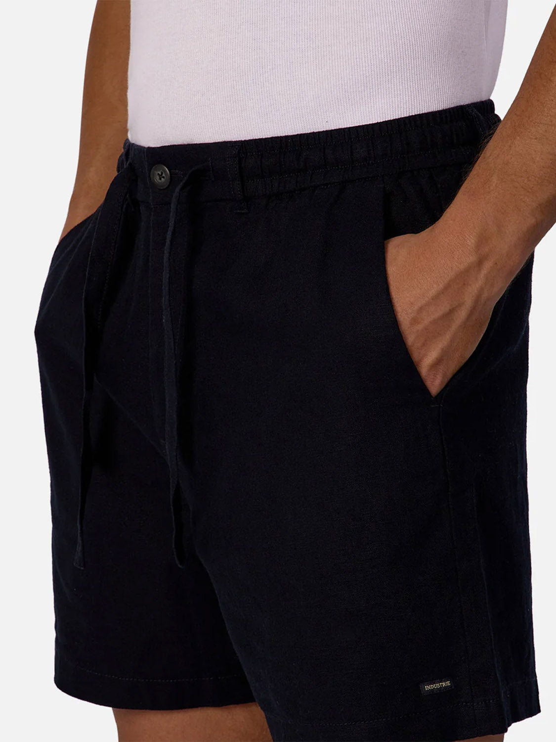 A person wearing a white tank top and Industrie Palma Linen Shorts in black, with hands in the pockets, shown from waist to mid-thigh.