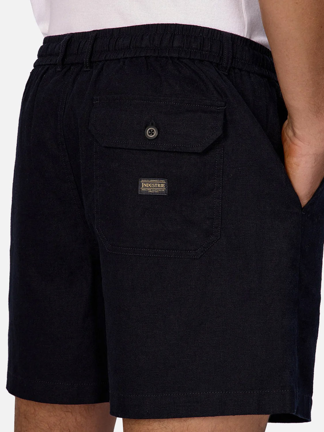 A close-up of a person wearing Industrie’s Palma Linen Short—black men's casual shorts with an elastic waistband, flap pocket with button, small Industrie label above the pocket, and the person's hand in the side pocket.