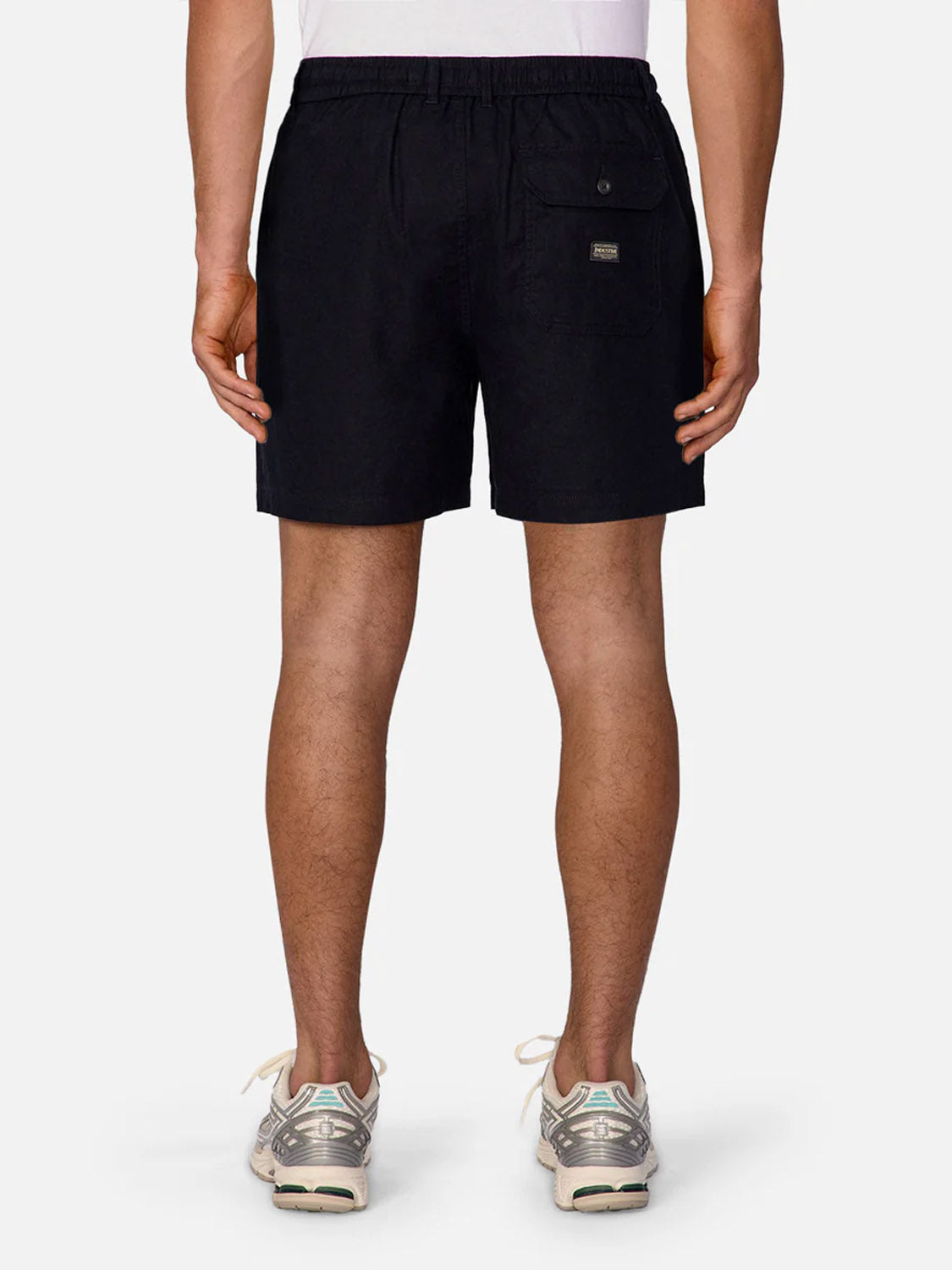 A person stands facing away, wearing a white shirt, Industrie Palma Linen Short in black with a back pocket, and gray sneakers with white soles. The background is plain and light-colored.