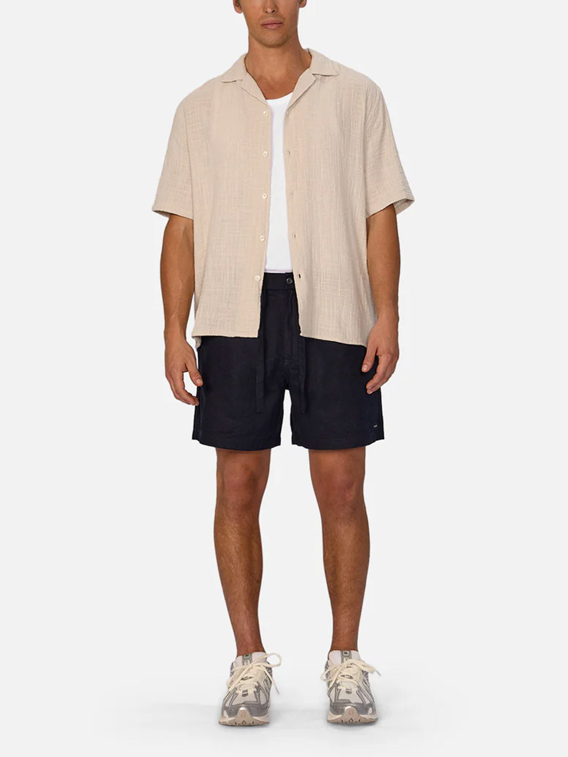 A man stands facing forward against a plain white background, wearing Industrie’s Palma Linen Short in black, paired with a beige short-sleeve button-up over a white T-shirt and beige athletic sneakers.