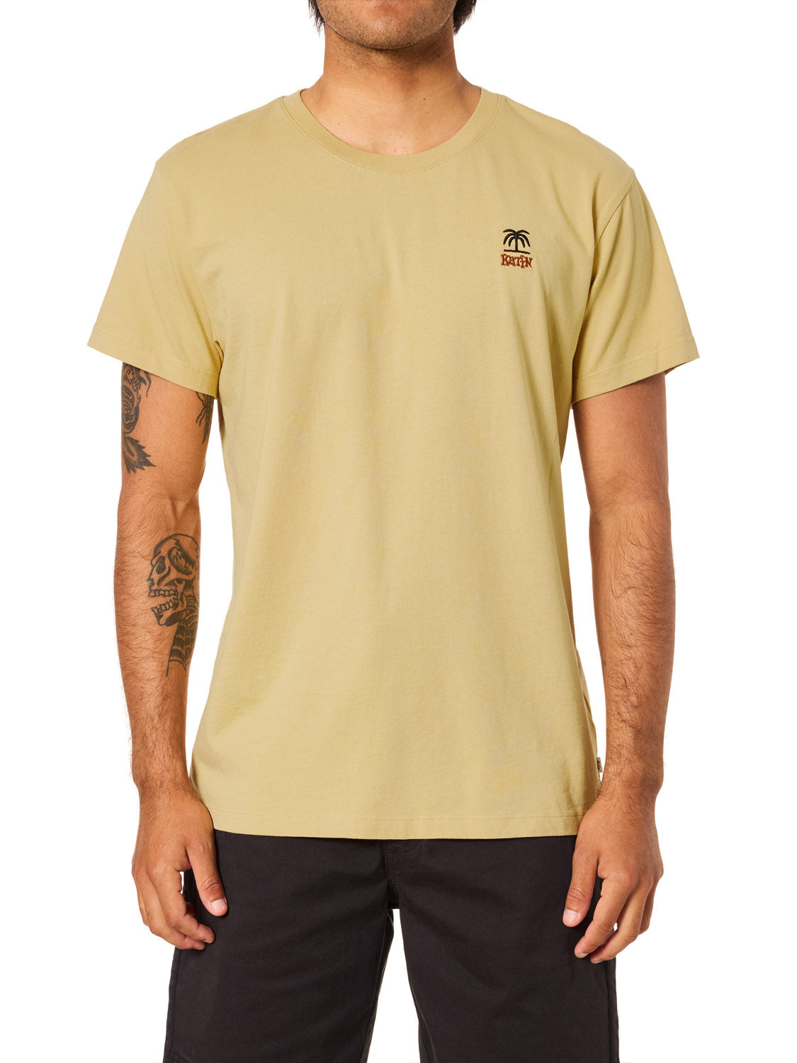 A person wears the Katin K-Palm Embroidered Tee, a light tan organic cotton short-sleeve shirt with a small palm tree and logo on the left chest by Katin, paired with black pants, revealing tattoos on their forearms.