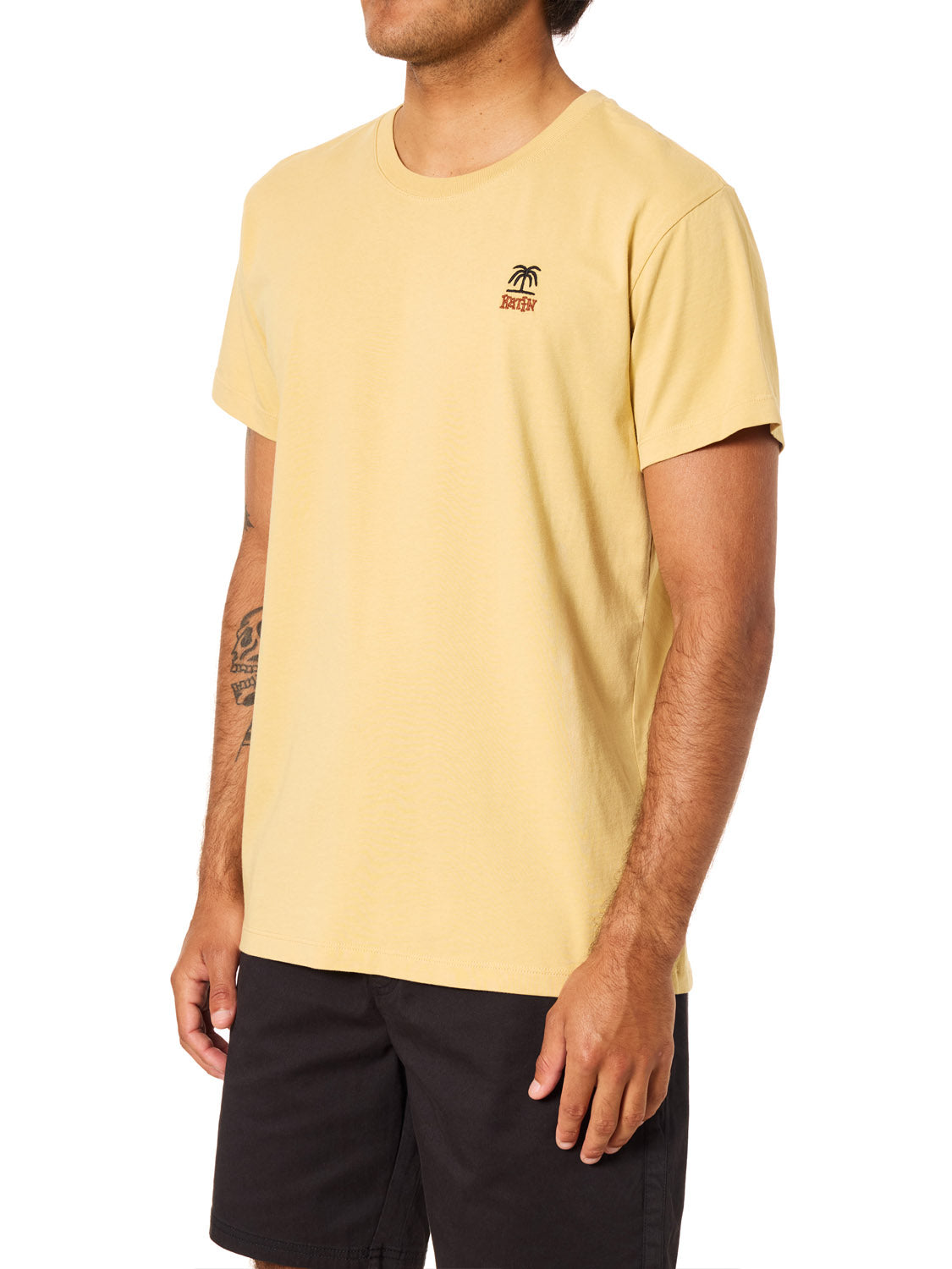 A person wears the Katin K-Palm Embroidered Tee by Katin, a light yellow organic cotton shirt featuring a small palm tree graphic on the chest, paired with black shorts, standing against a white background with face not visible.