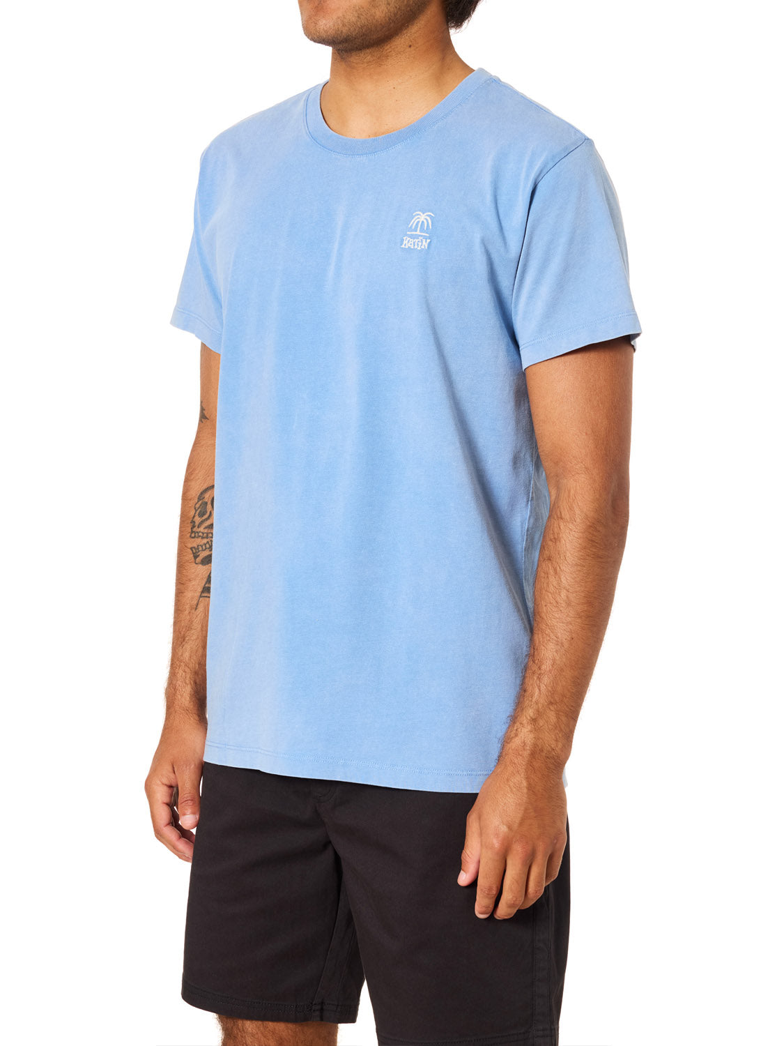 A man wears the Katin K-Palm Embroidered Tee by Katin—light blue organic cotton with a small white chest logo—and black shorts, standing against a plain white background.
