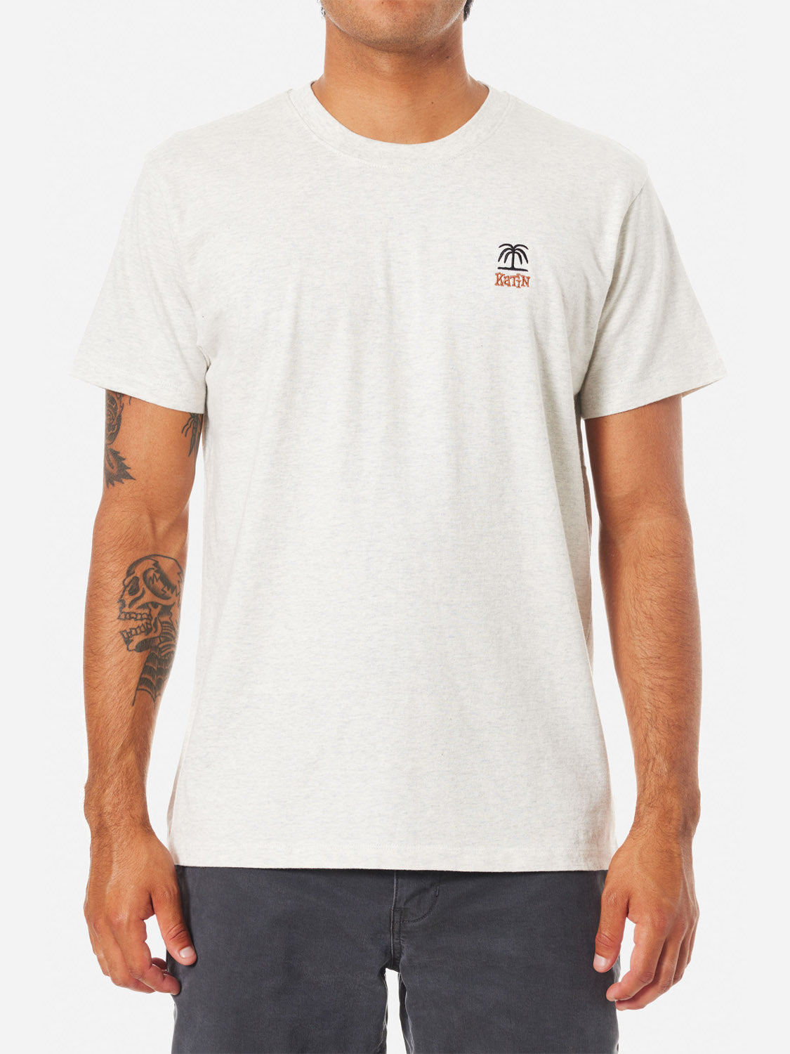 A man wears the light grey Katin Palm Embroidered Tee—organic cotton with a small palm tree and ROTTN logo on the chest—paired with dark shorts. Tattoos are visible on his right forearm as he stands against a plain white background.