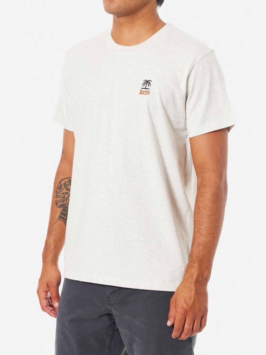 A man wears the Katin Palm Embroidered Tee—a short-sleeved, light beige organic cotton shirt with a small palm tree and Katin logo on the left chest—paired with dark pants and a visible right forearm tattoo for vintage surf style.