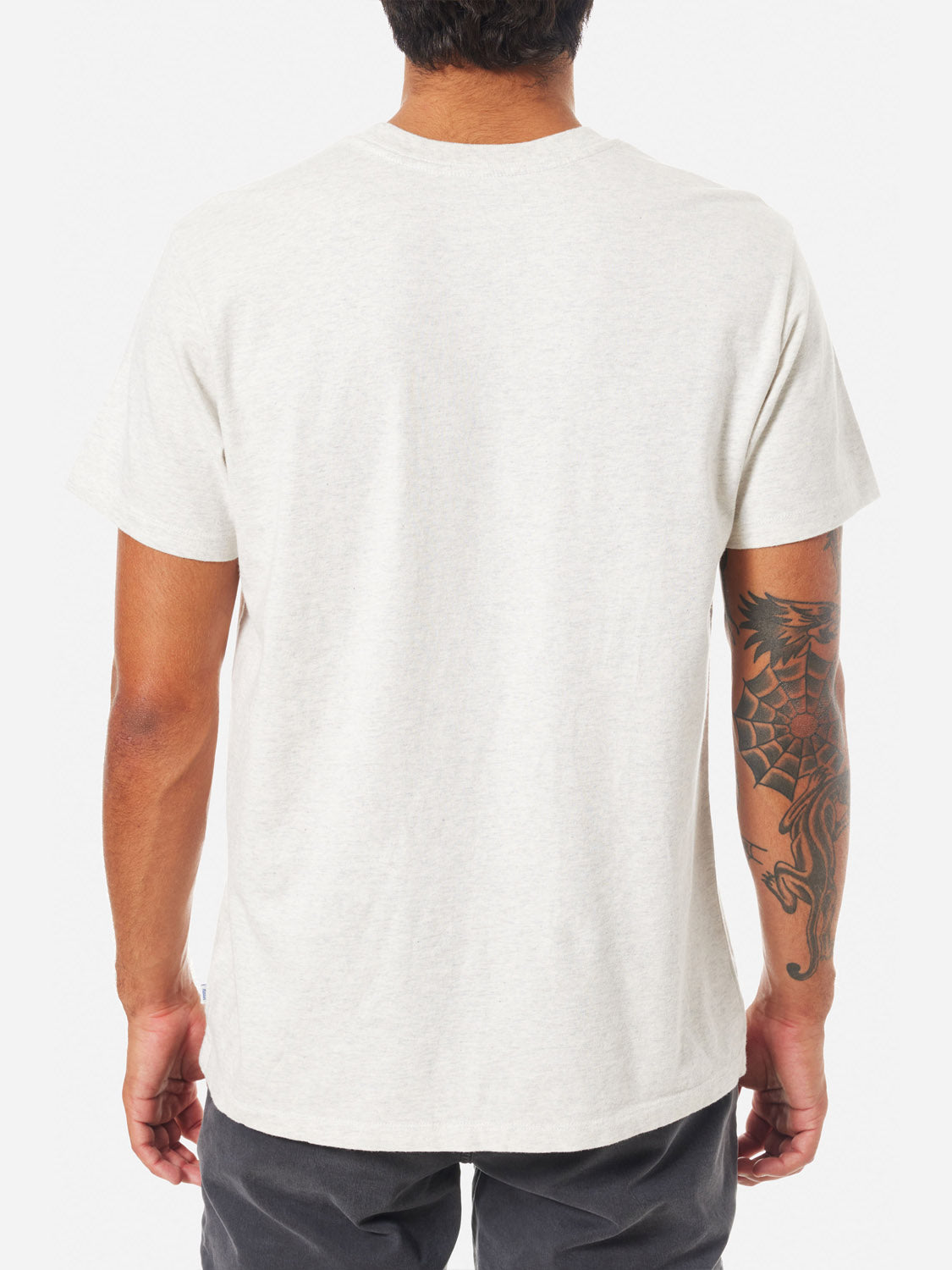 A person with short dark hair is seen from behind wearing the Katin Palm Embroidered Tee by Katin in light gray organic cotton, paired with dark pants. A large dragon tattoo covers their right arm.