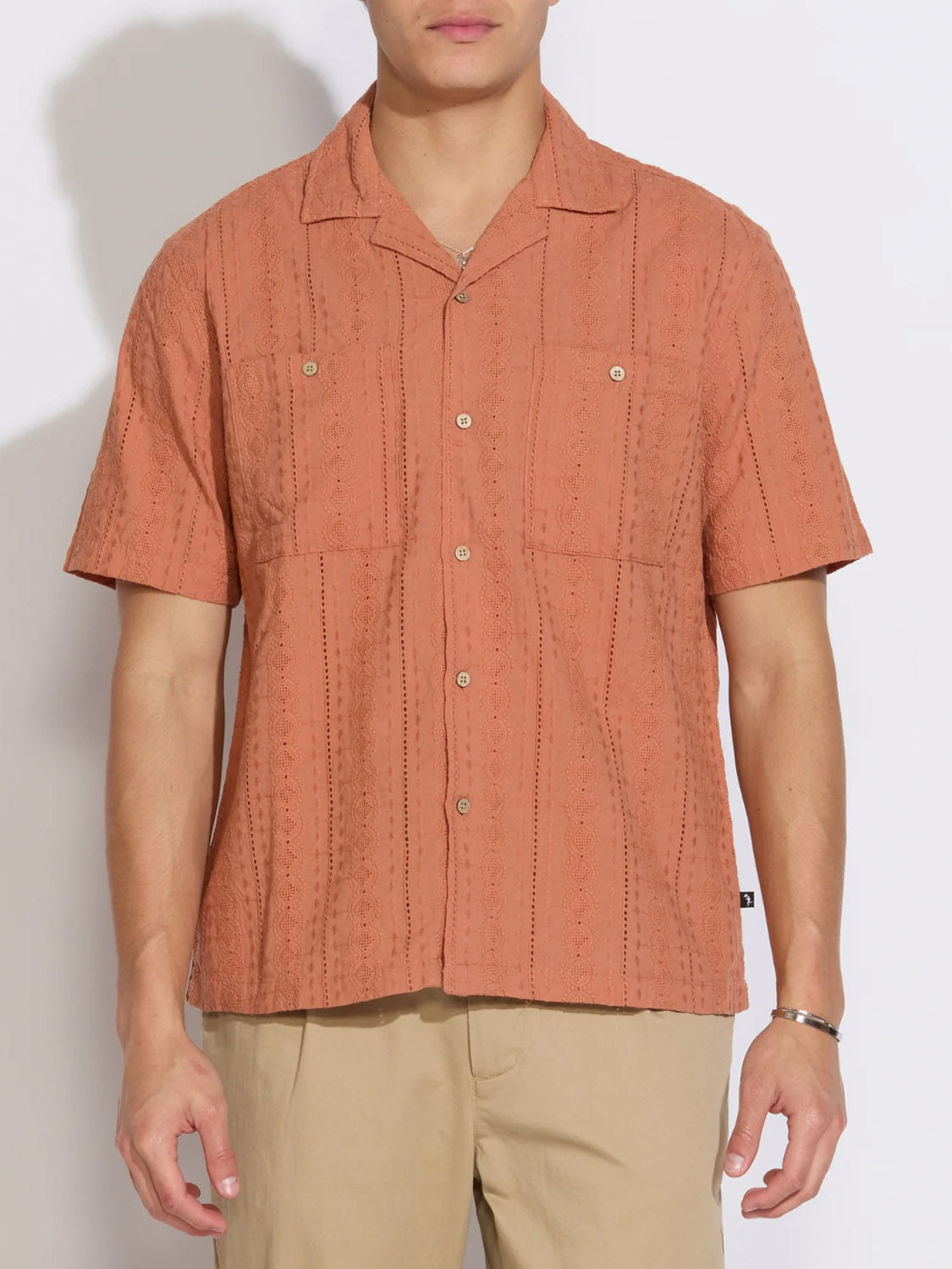 A person wears the Civil Society Palomino Relaxed Fit Pocket Resort Shirt—a short-sleeve, button-up textured rust-orange Cuban collar shirt with two chest pockets—paired with beige pants, standing against a plain white background.