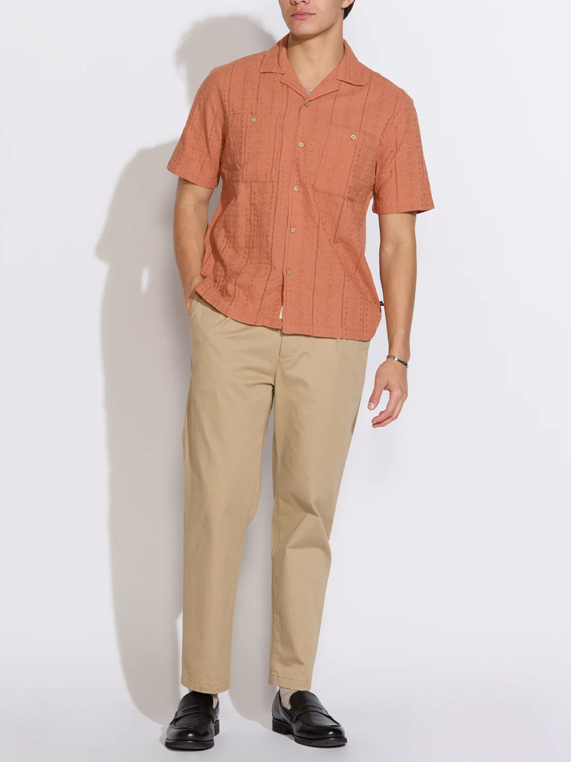 A man poses against a white background wearing the Palomino Relaxed Fit Pocket Resort Shirt by Civil Society in rust, paired with beige pants, black loafers, and a watch, with one hand in his pocket.
