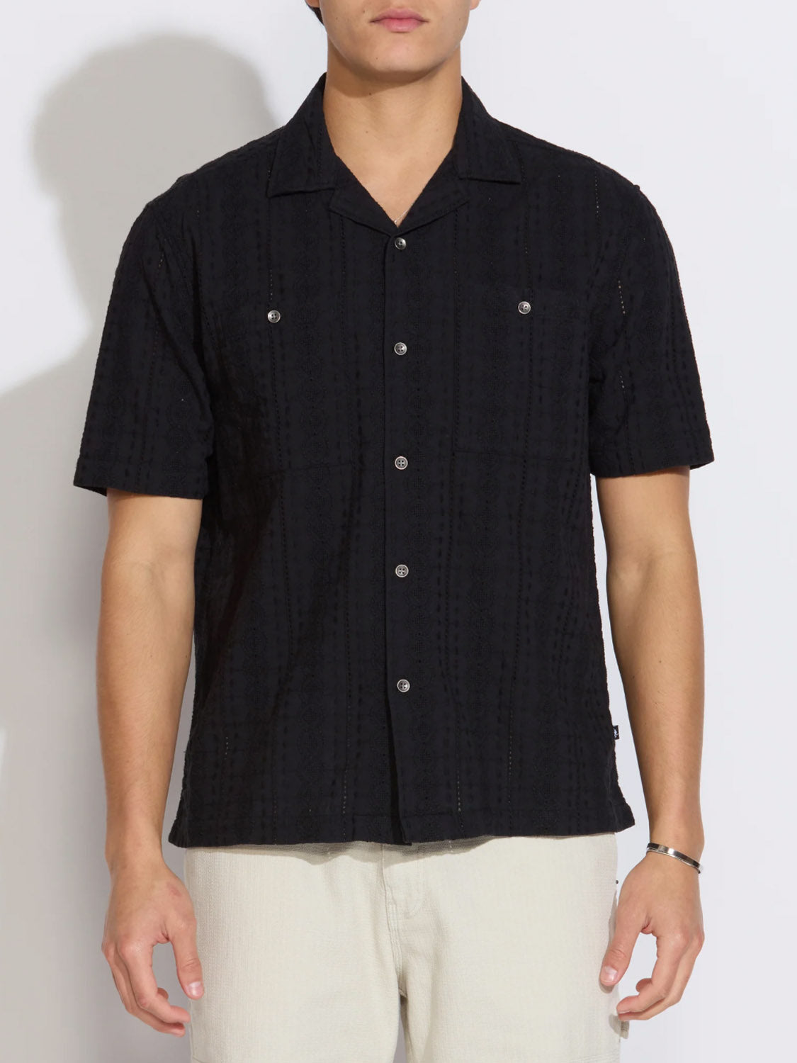 A person wears the Civil Society Palomino Relaxed Fit Pocket Resort Shirt—a black, short-sleeve Cuban collar shirt with a subtle textured pattern—paired with light-colored pants, standing against a plain white background.
