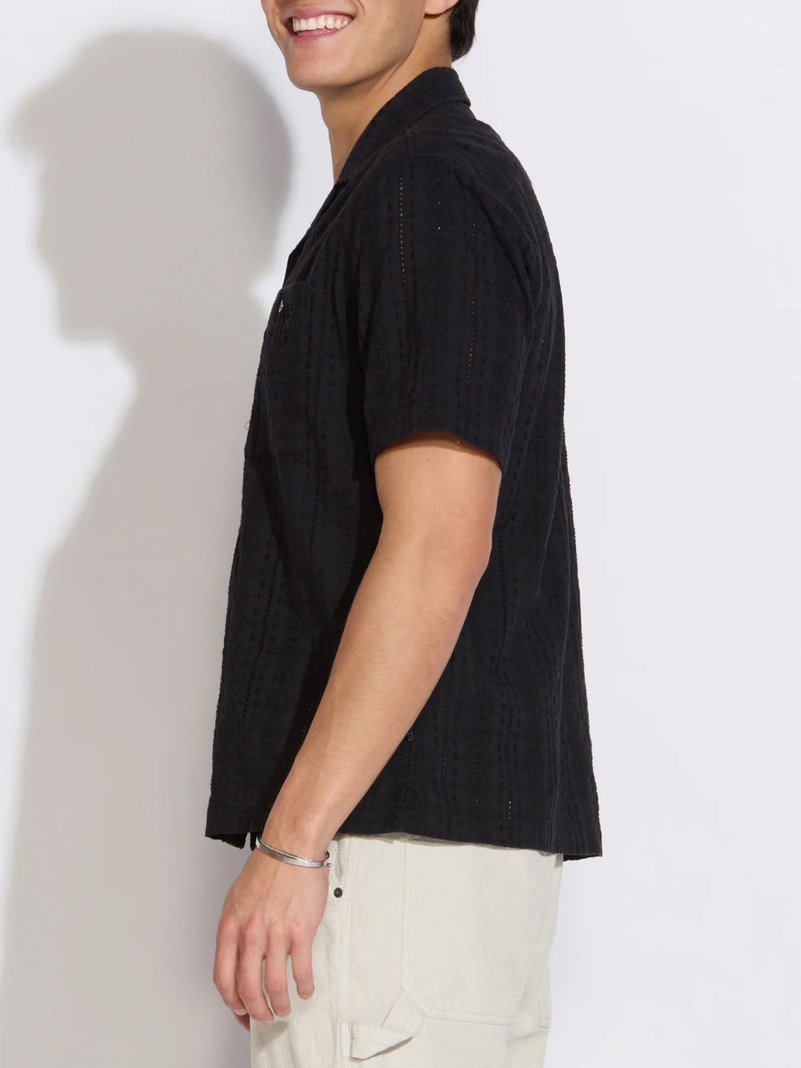 A person stands sideways, smiling, in the Civil Society Palomino Relaxed Fit Pocket Resort Shirt (short-sleeve, black) with light-colored pants against a plain white background.