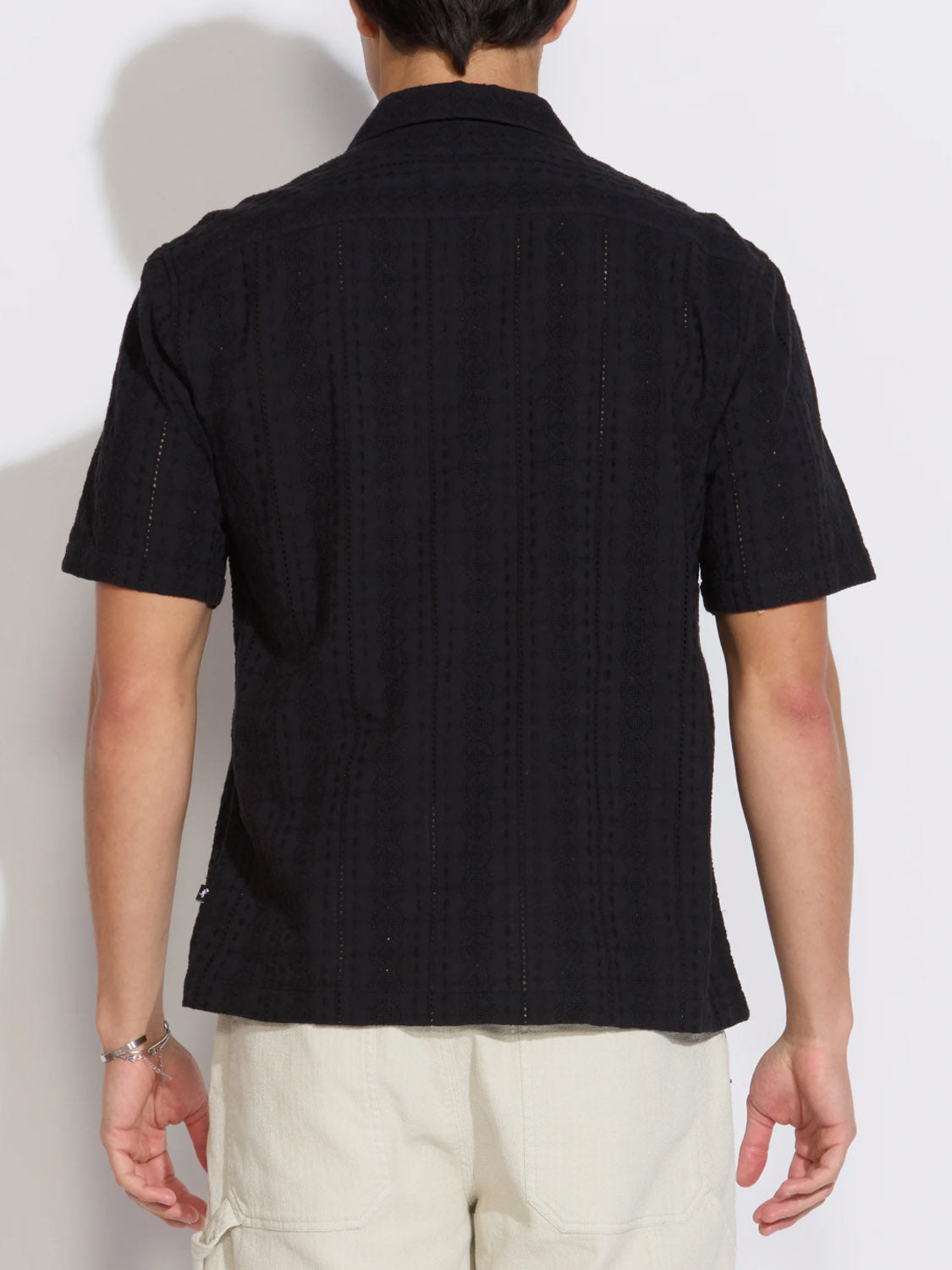A person faces away from the camera, wearing the Civil Society Palomino Relaxed Fit Pocket Resort Shirt in black paired with light shorts. The shirt features a Cuban collar and subtle textured pattern. The background is plain and well-lit.