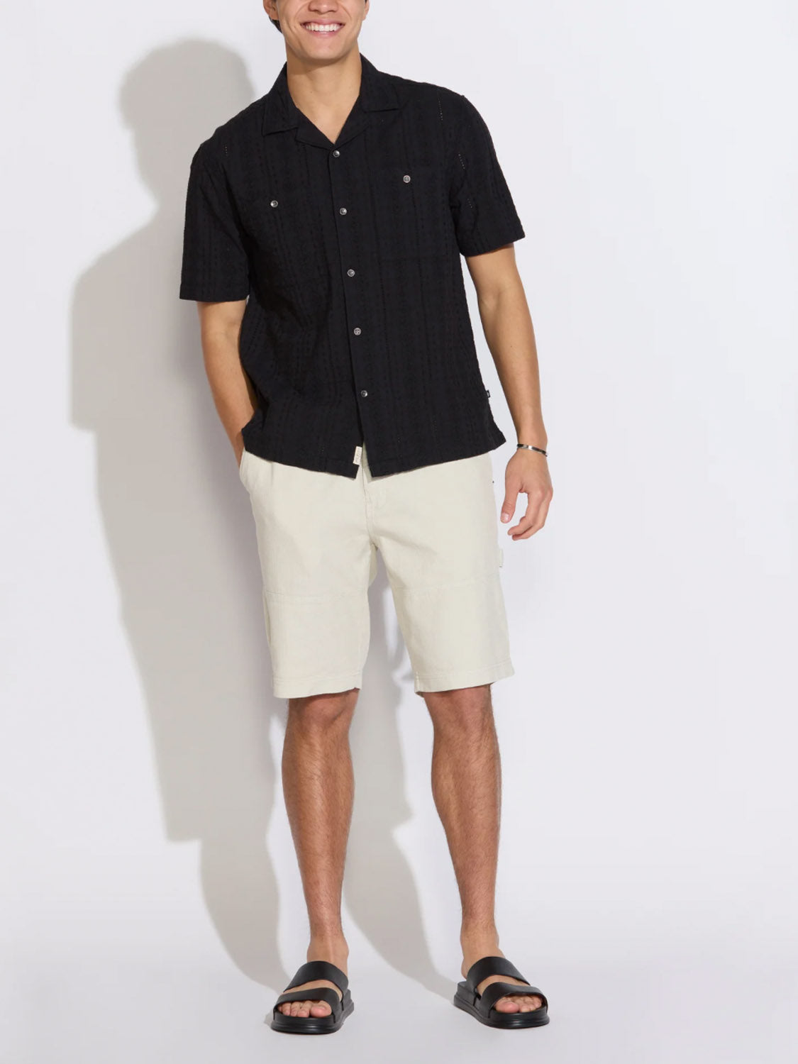 A man stands against a plain white background wearing the Civil Society Palomino Relaxed Fit Pocket Resort Shirt, light beige shorts, black sandals, and a bracelet, hands in his shorts pockets and looking relaxed.
