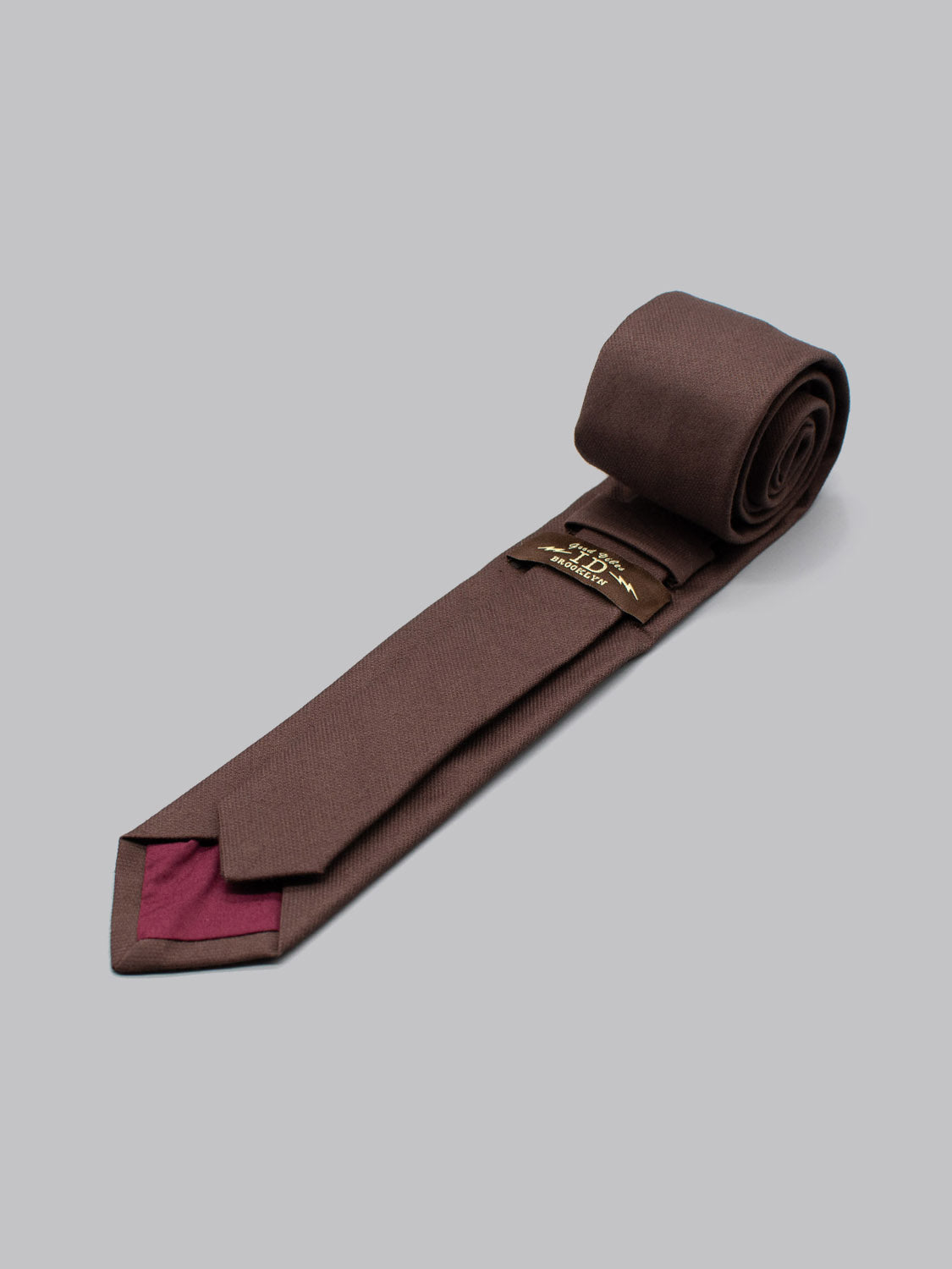 The ID Menswear ID Cotton Tie Paris is a brown handmade cotton tie with subtle texture and a maroon accent on the underside, shown partially unrolled on a light gray background—a refined business formal choice.