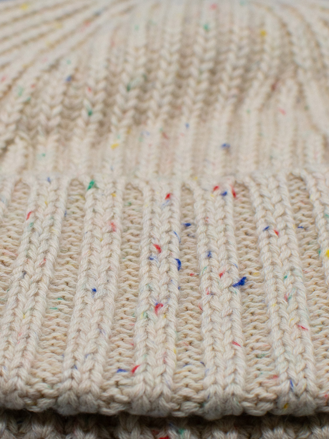 Close-up of a beige, ribbed-knit fabric resembling an ID Menswear Paro Speckled Cotton Fishermans Beanie. It boasts a timeless design with colorful specks of red, blue, and green yarn interspersed throughout, creating a subtle pattern.