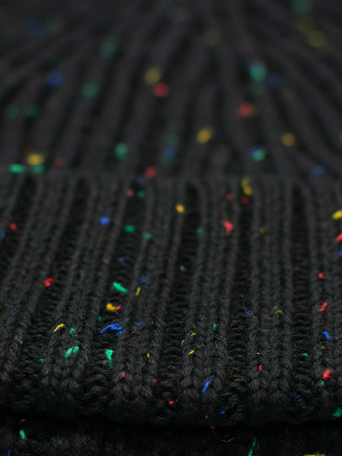 Close-up of black ribbed knit fabric made from speckled yarn in red, blue, yellow, and green hues—a timeless design resembling the Paro Speckled Cotton Fishermans Beanie by ID Menswear.