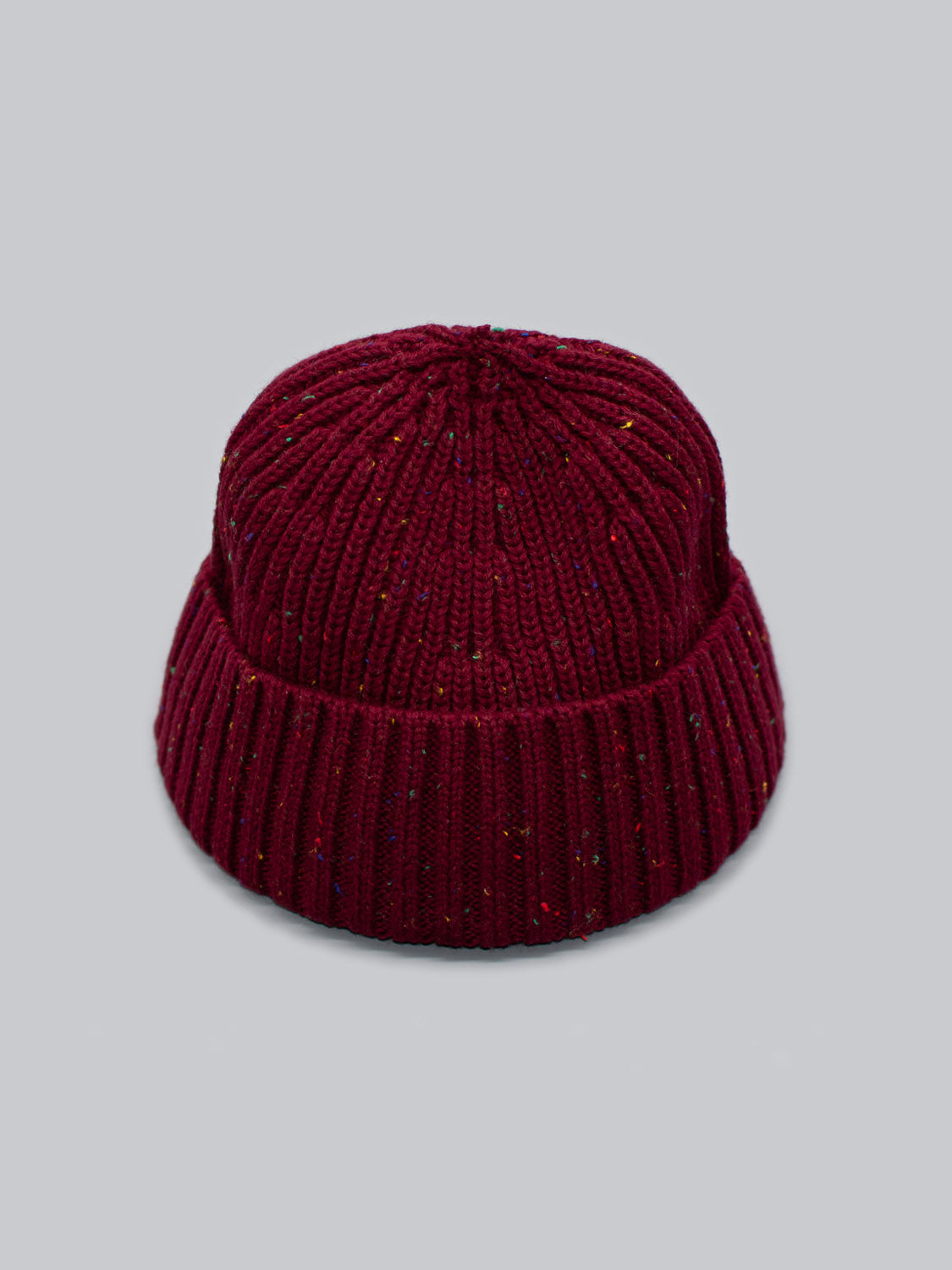 The Paro Speckled Cotton Fishermans Beanie from ID Menswear, in deep red speckled yarn with a folded brim, is displayed on a light gray background, highlighting its timeless design.