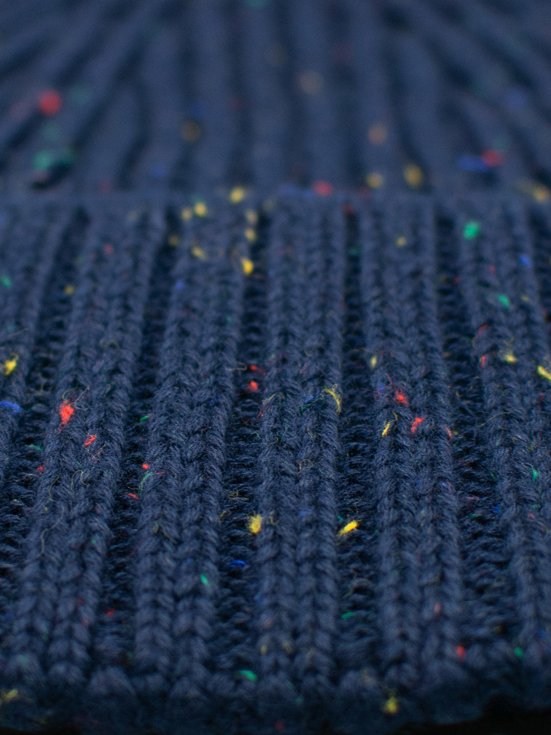 Close-up of a knitted navy-blue fabric with ribbed texture and scattered colorful specks in yellow, red, and blue. This yarn pattern reflects fine craftsmanship and cozy appeal, similar to the timeless design in ID Menswears Paro Speckled Cotton Fishermans Beanie.